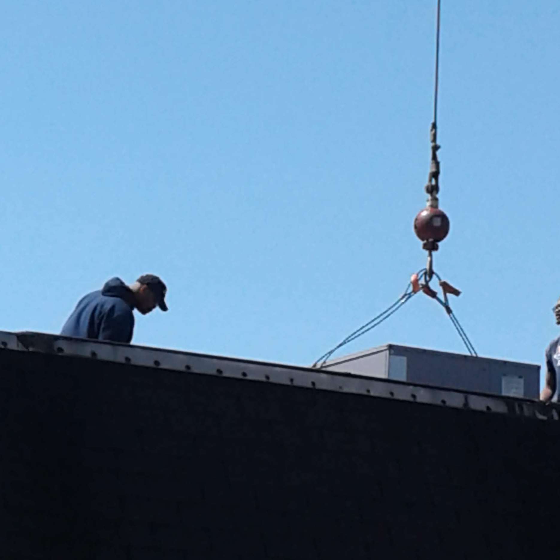 Workers installing an HVAC unit on a roof with a crane on a clear, sunny day.