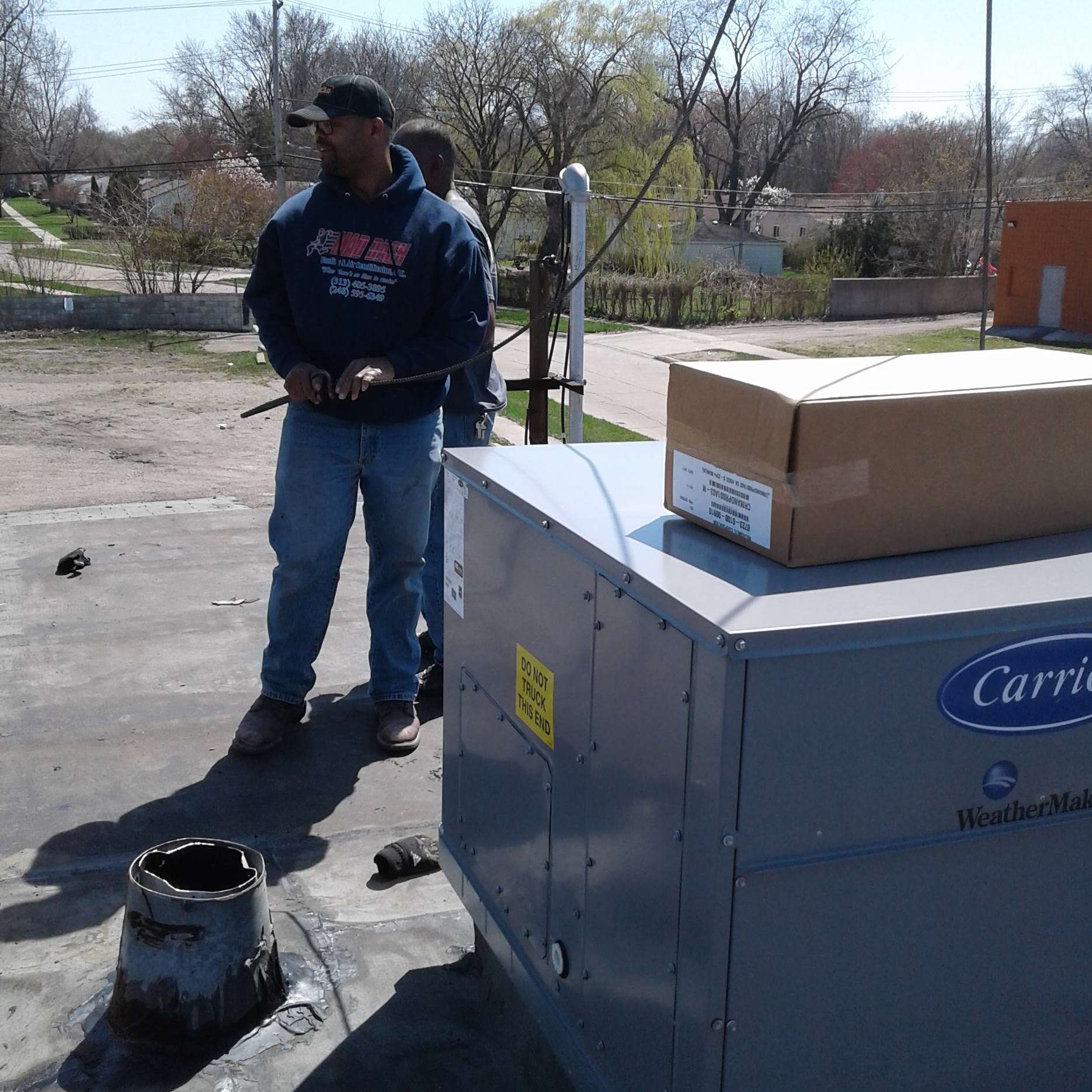 Man in sweatshirt and jeans on rooftop near Carrier HVAC unit. Cardboard box on top.