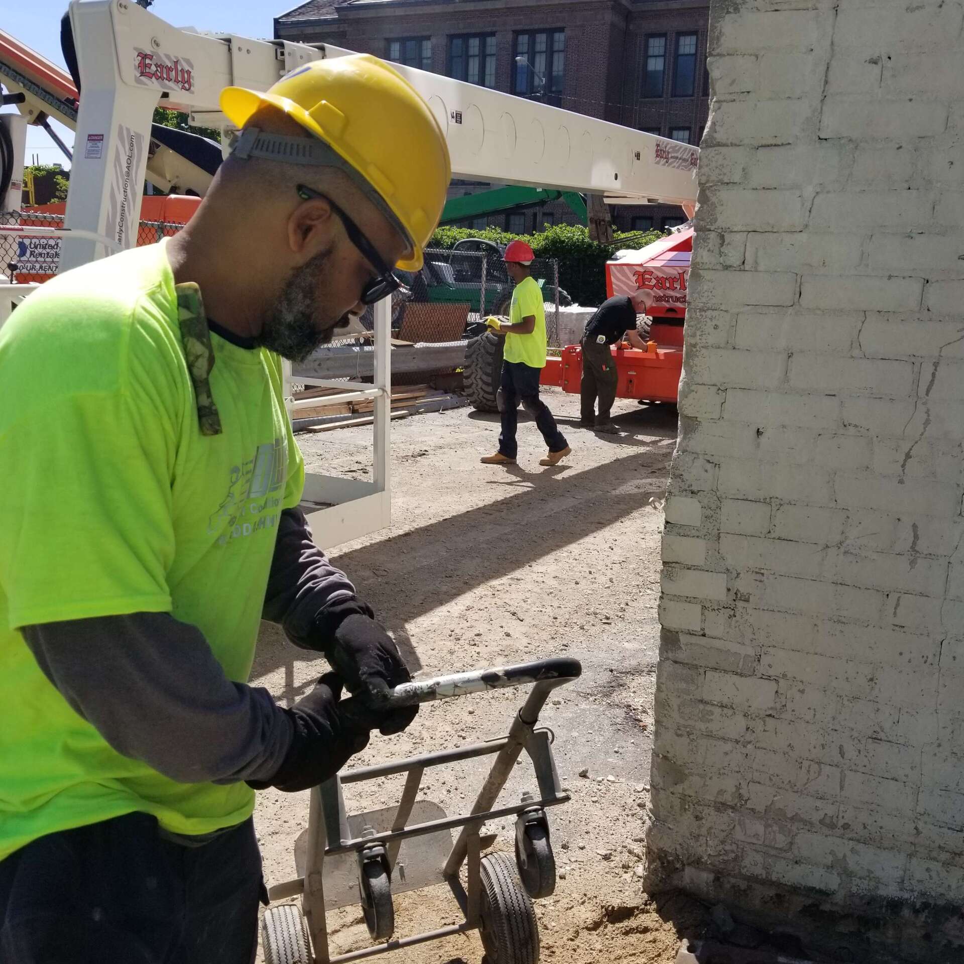 Construction worker in yellow safety gear operates a tool near a white brick wall.