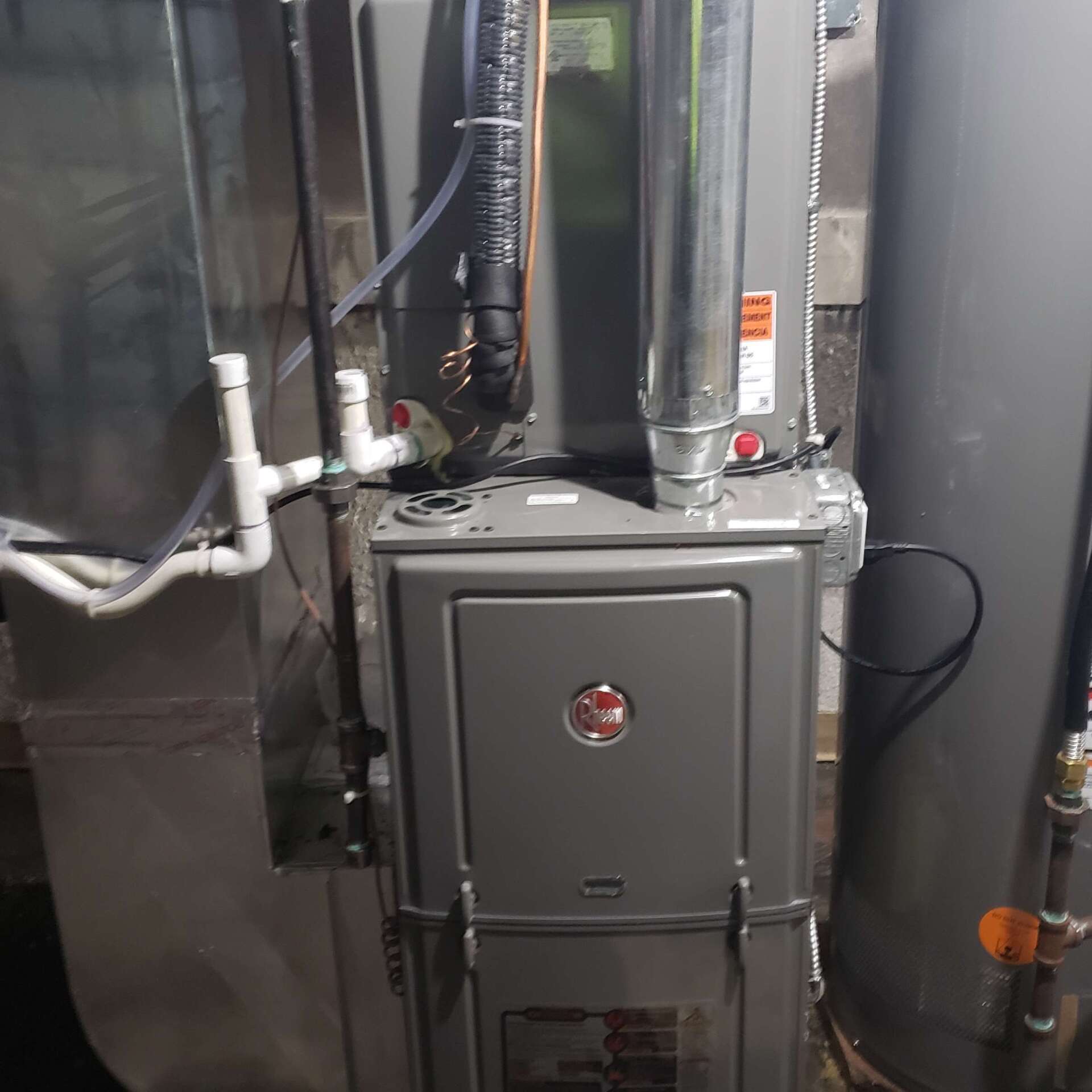 A gray furnace and water heater in a utility room with various pipes and ducts.