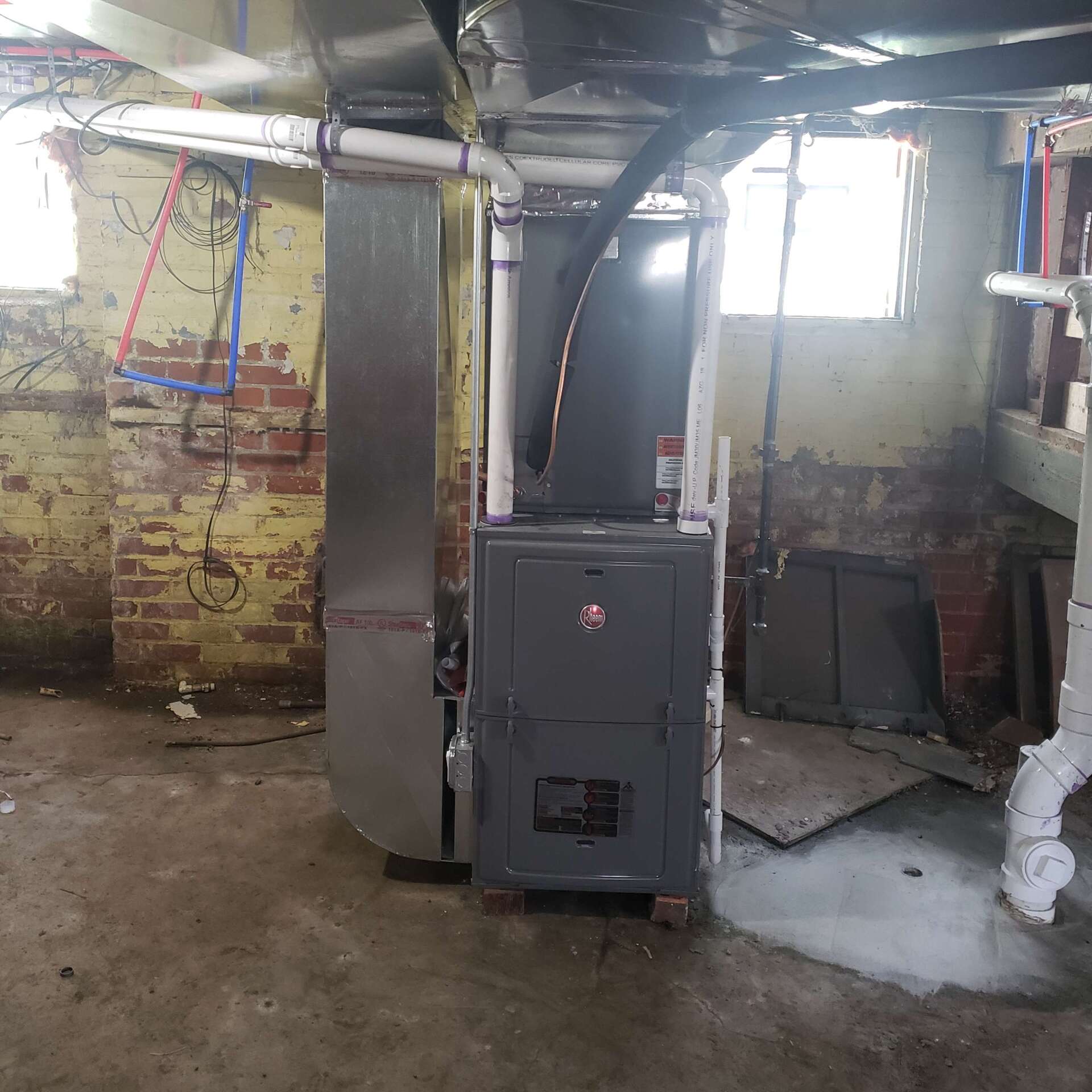 A gray furnace in a basement with pipes and a brick wall.