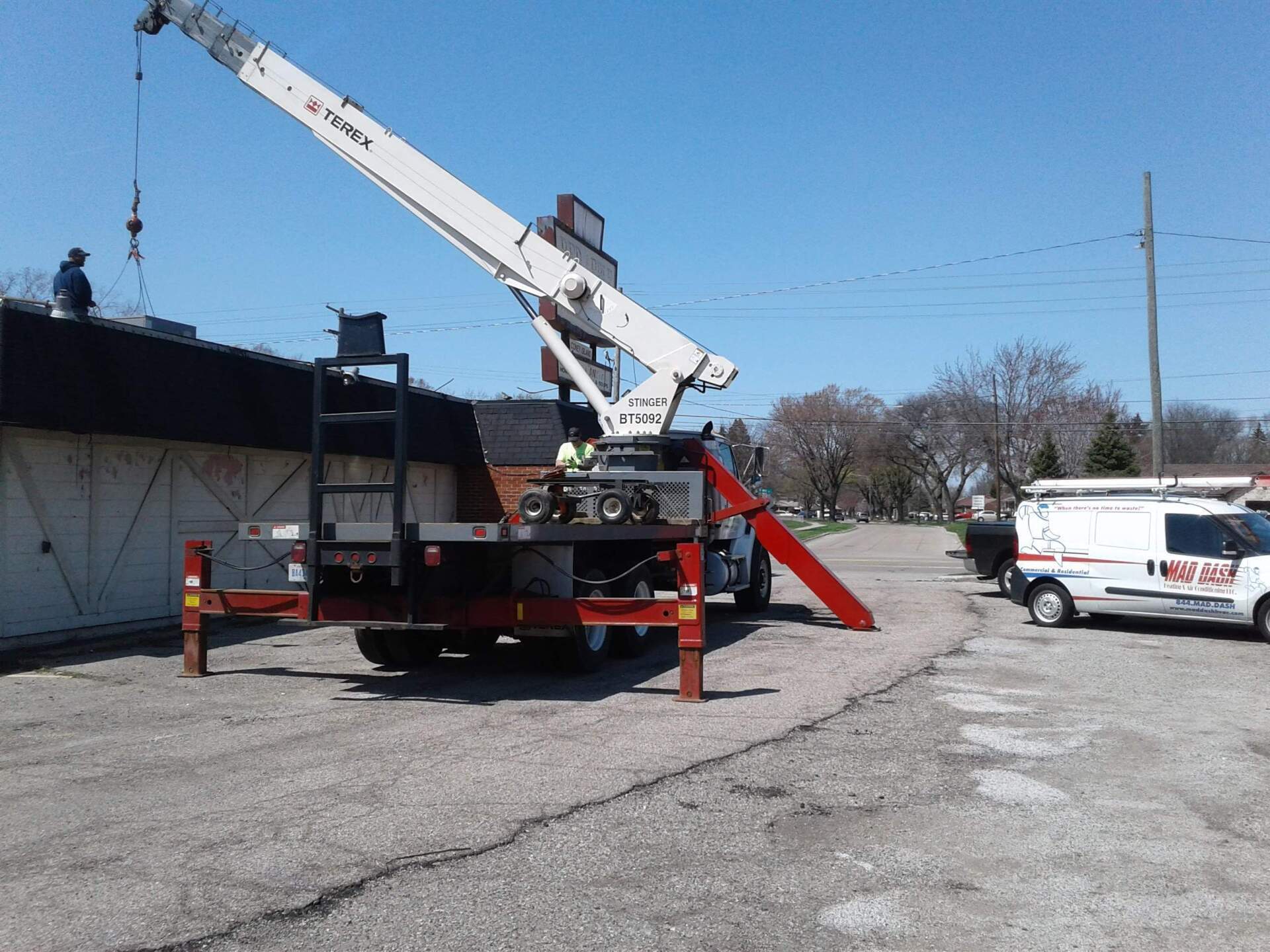 A crane lifts something onto a dark rooftop. A white van is parked nearby on a sunny day.