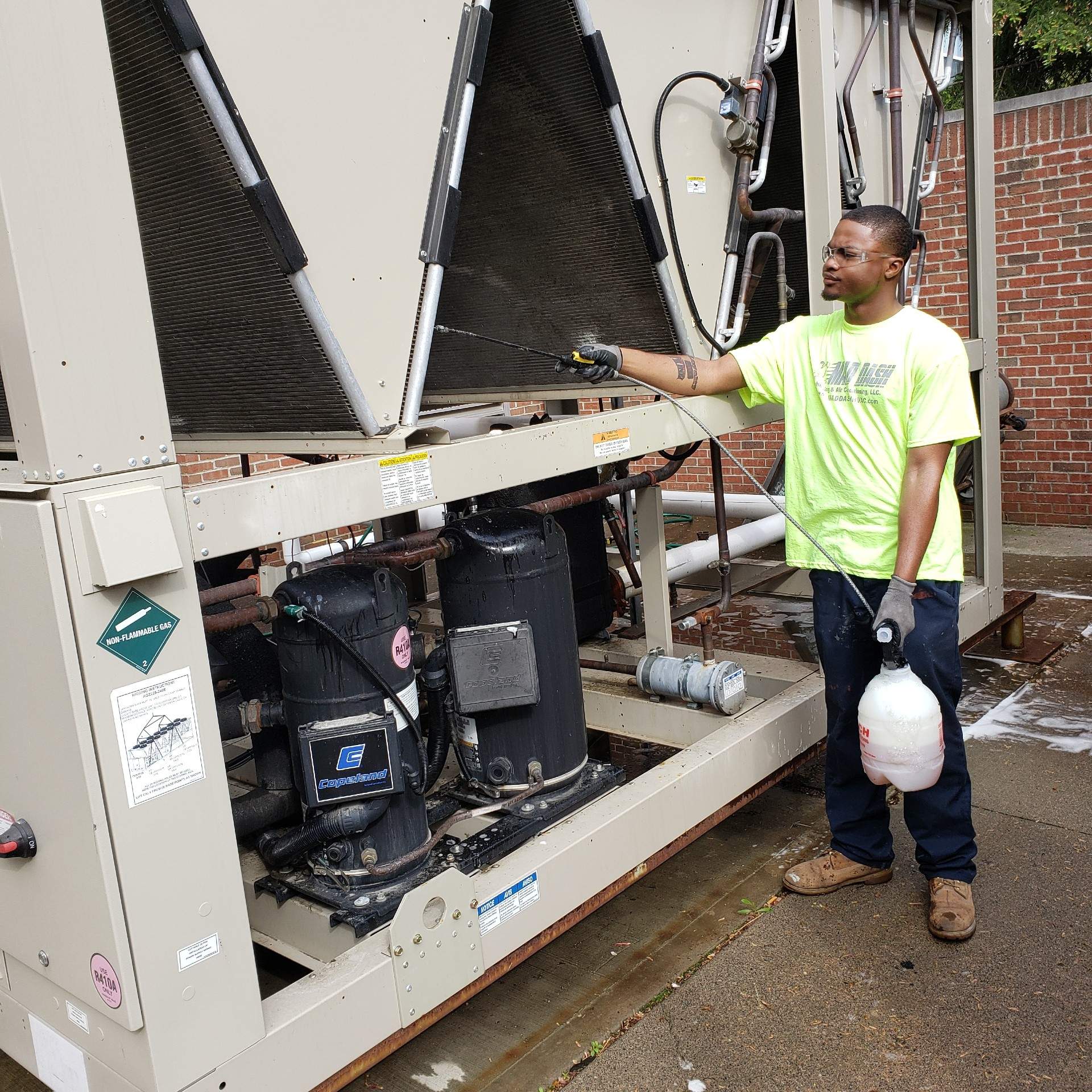 Man in neon shirt spraying an industrial air conditioning unit outside a brick building.