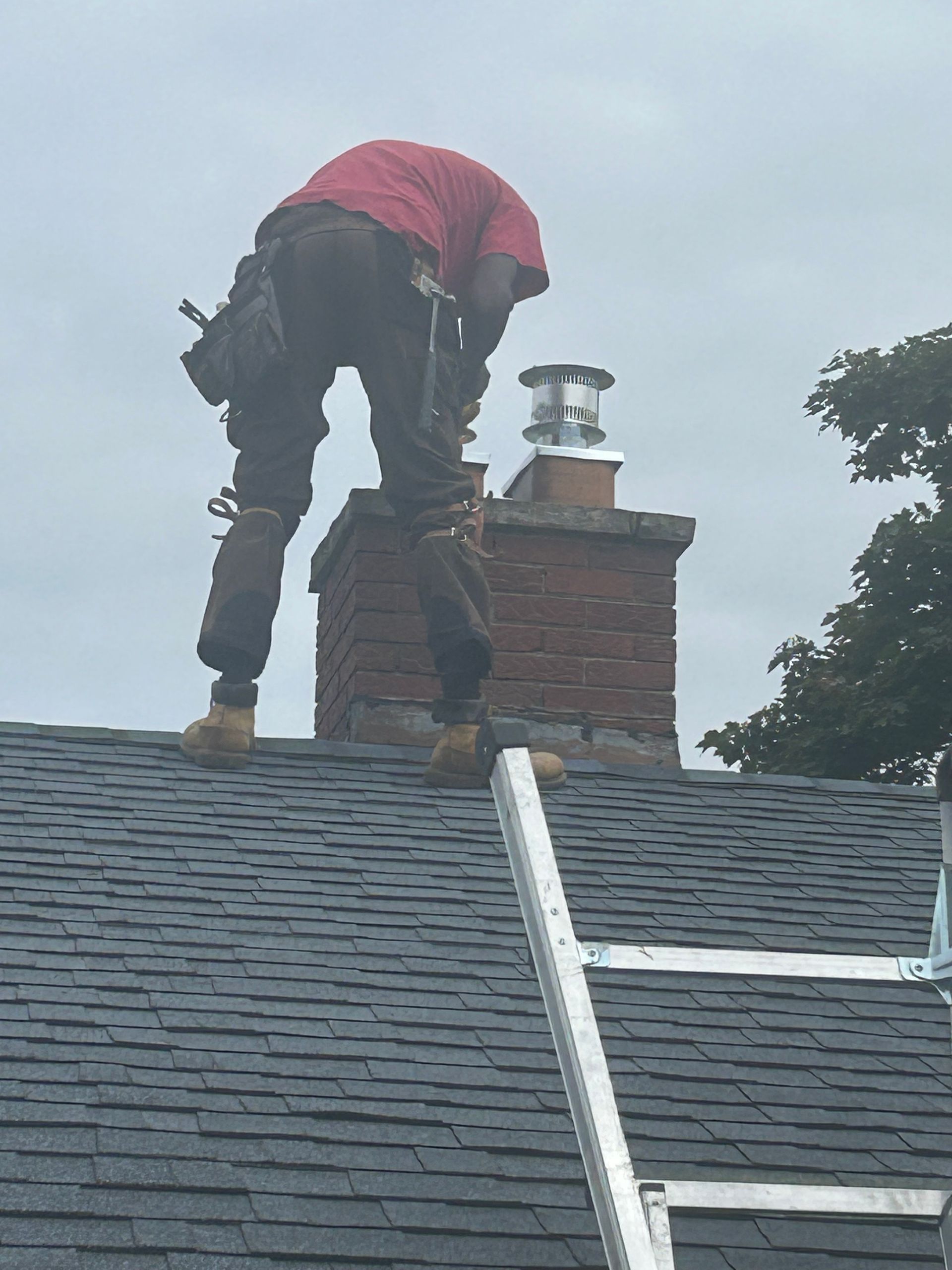 Roofer on a roof by a chimney, wearing red shirt and brown pants.