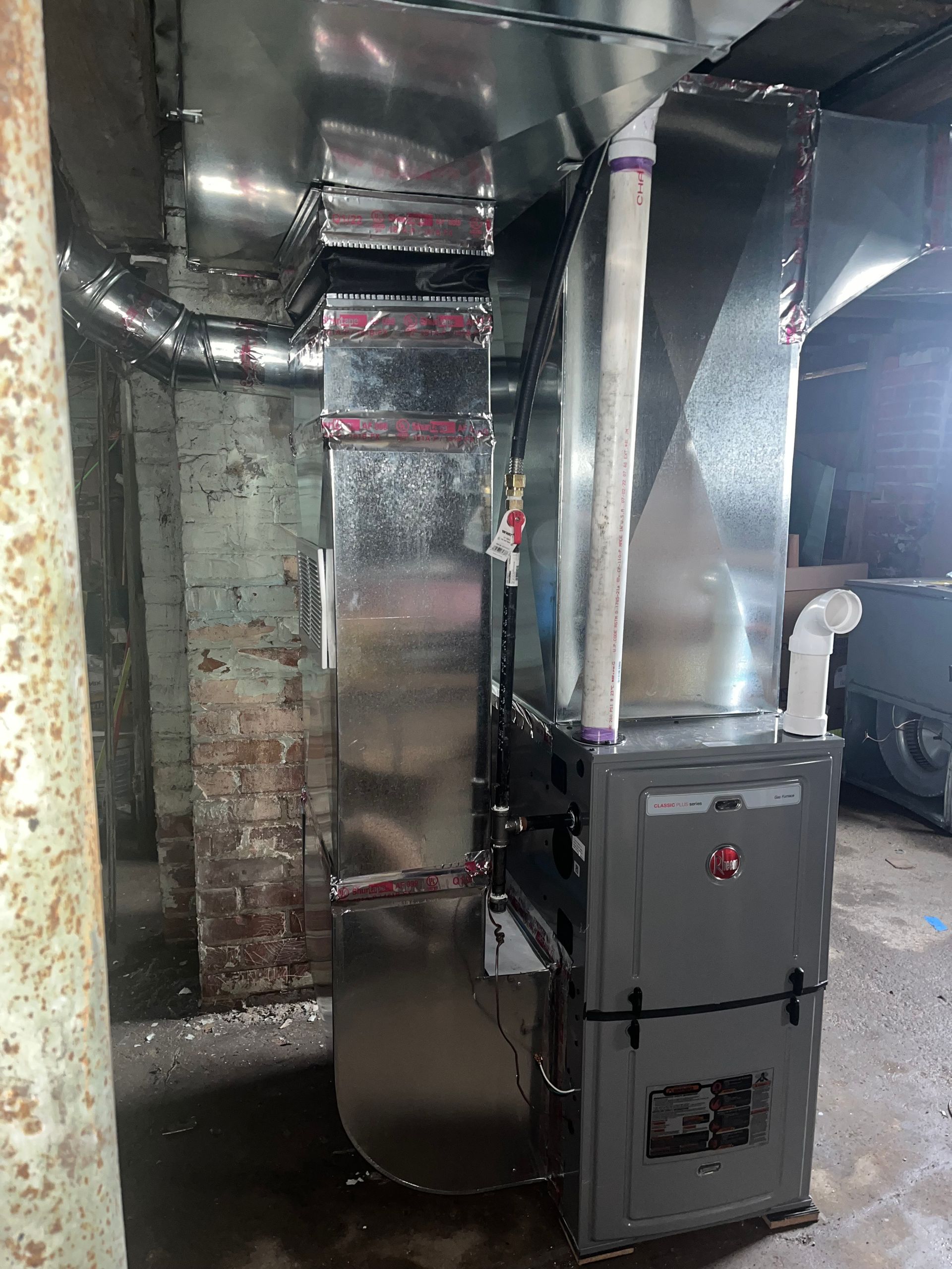 A new, gray Rheem furnace with silver ductwork installed in a brick-walled basement.
