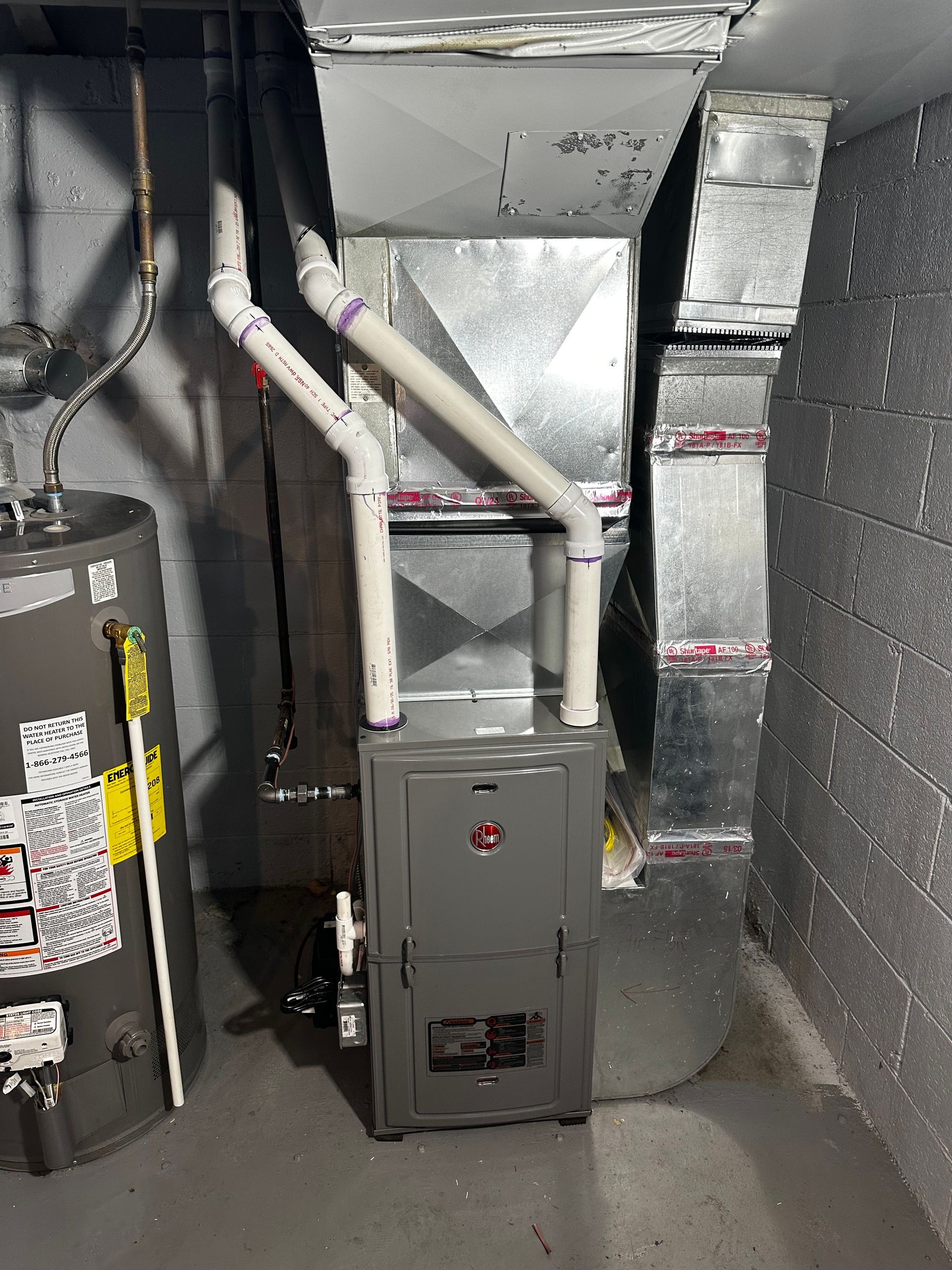 Furnace, water heater, and ductwork in a basement setting. Gray furnace, white pipes, gray ductwork, on a concrete floor.