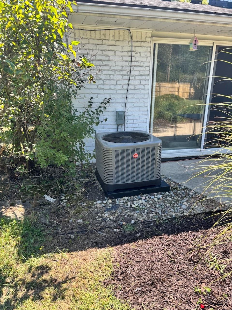 Outdoor air conditioning unit beside a house with a sliding glass door and brick wall, surrounded by mulch and greenery.