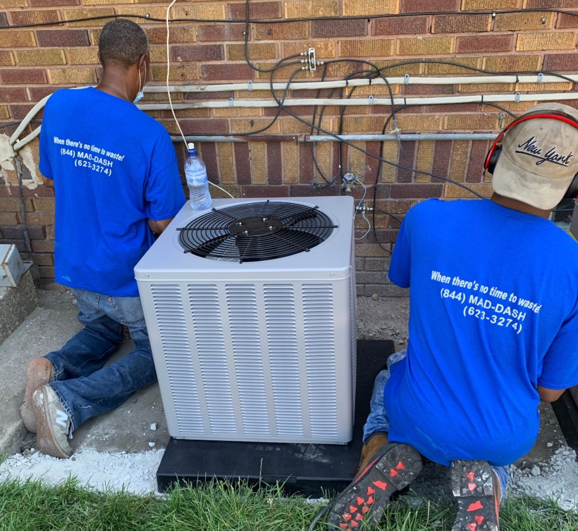 Two people in blue shirts installing an AC unit outside a brick building.