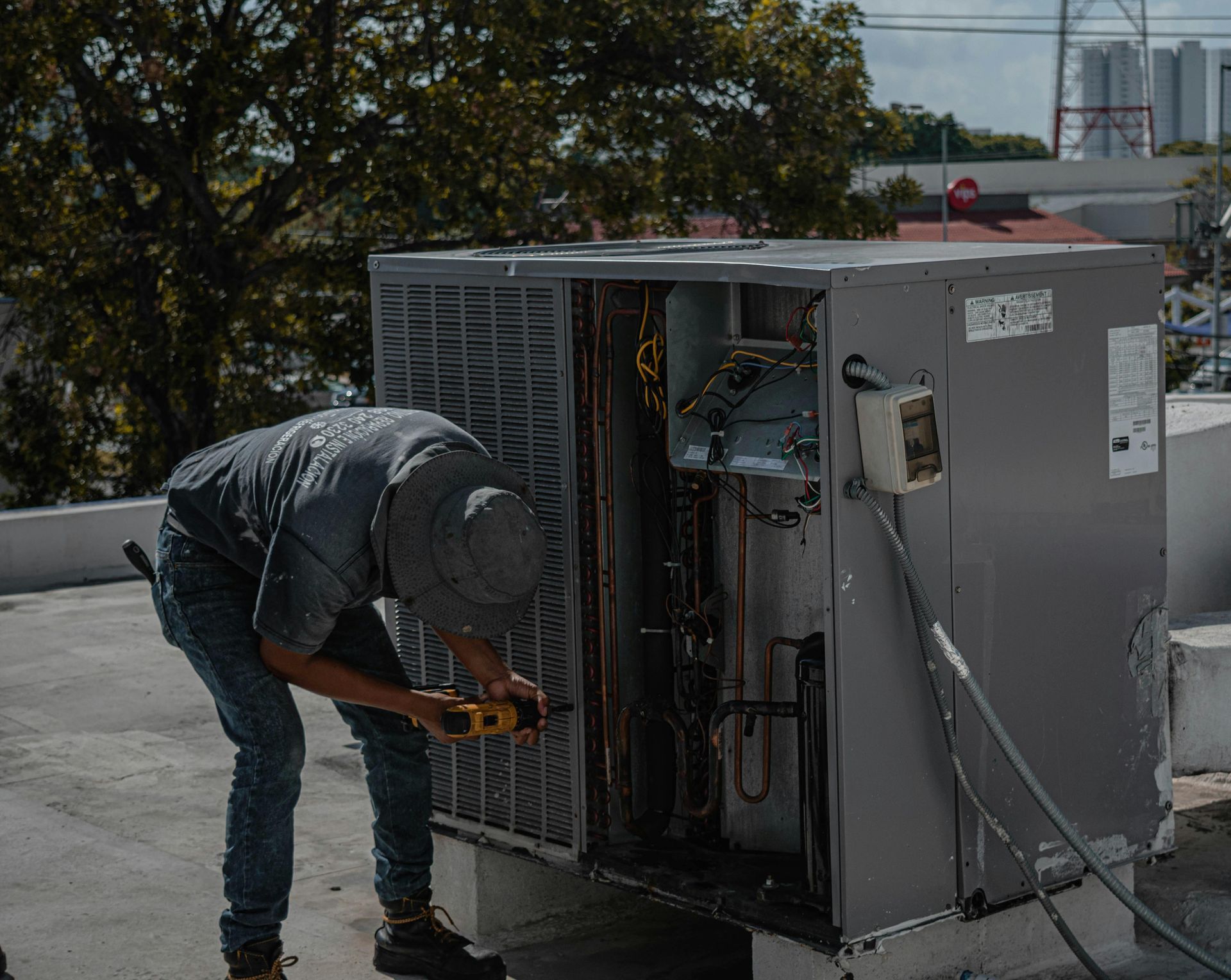 HVAC technician repairs an air conditioner on a rooftop. He wears a hat and jeans, using a tool.