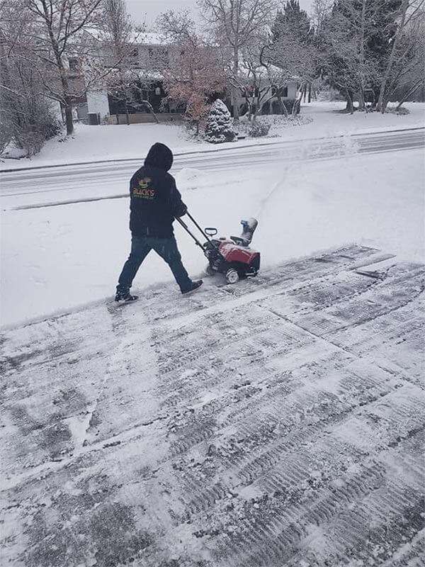 A man is pushing a snow blower down a snowy sidewalk.