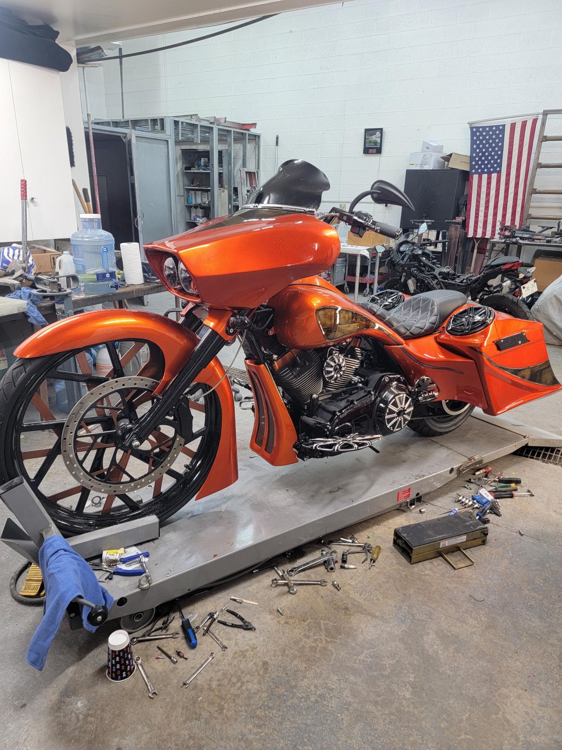 An orange custom motorcycle on a lift in a workshop, with an American flag hanging in the background.