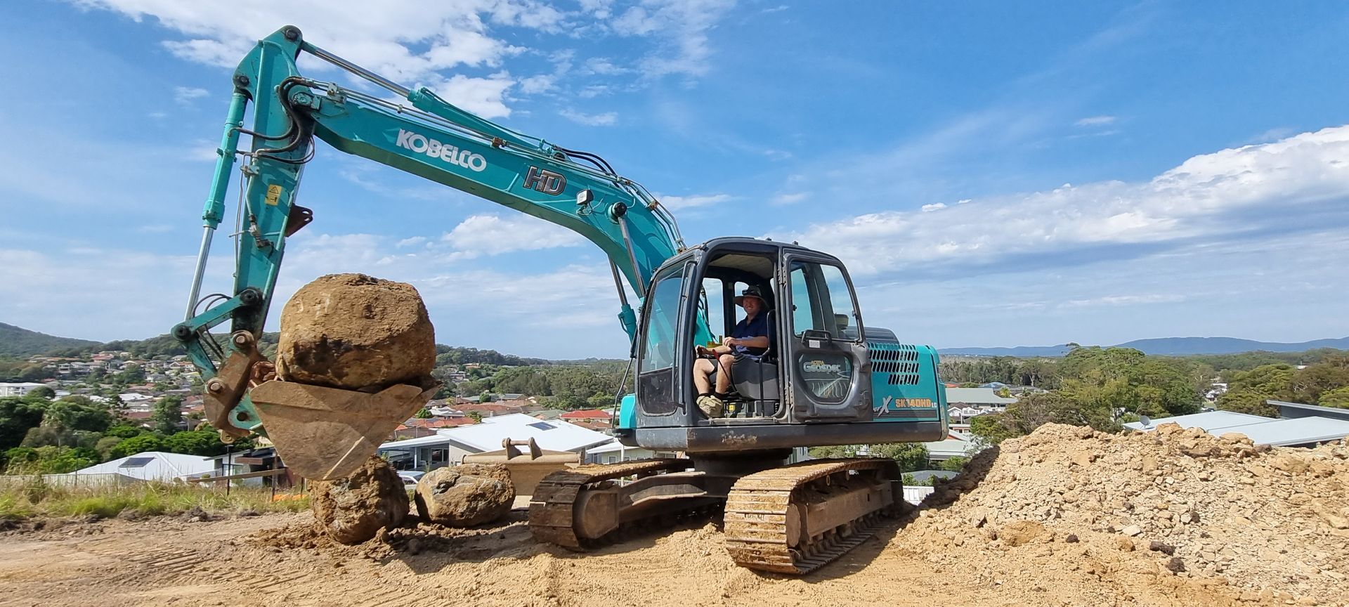 Excavator, blue and gray, scoops up a large chunk of dirt under a blue sky, construction site.
