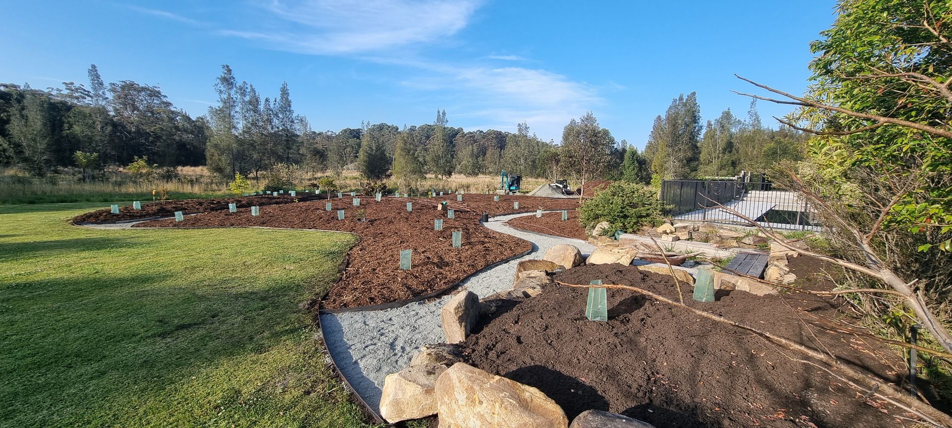 Garden with mulch, gravel path, and newly planted trees. Green lawn and blue sky in background.