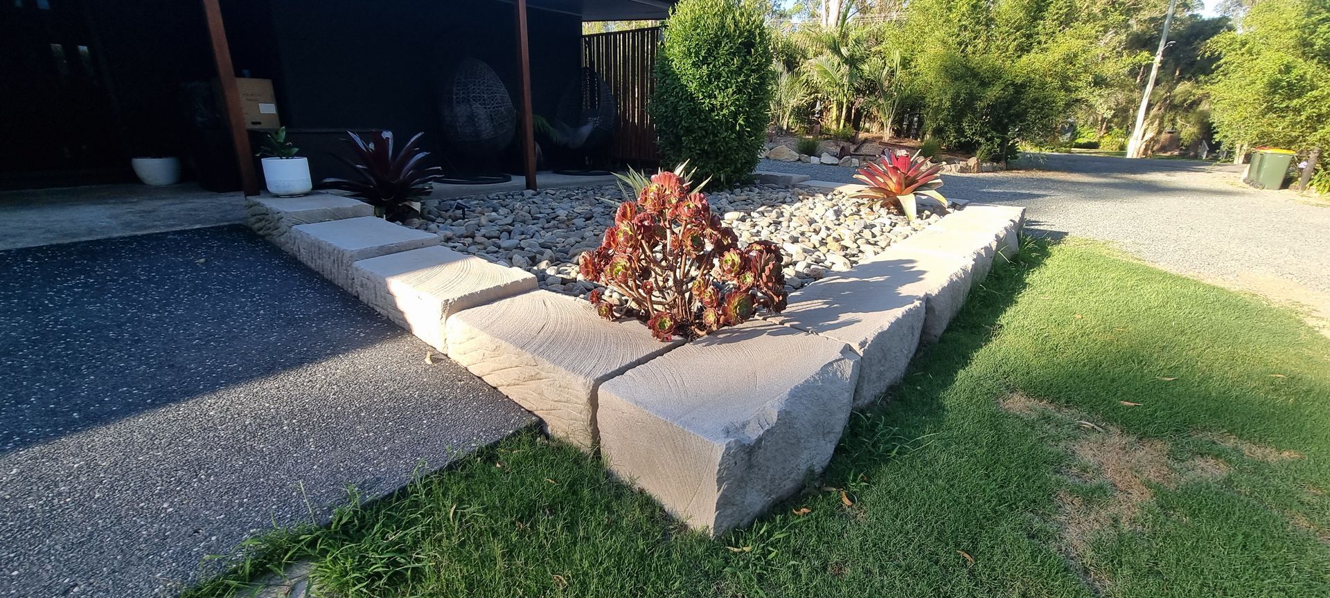 A rock garden with plants framed by stone blocks, beside a paved driveway and green lawn.