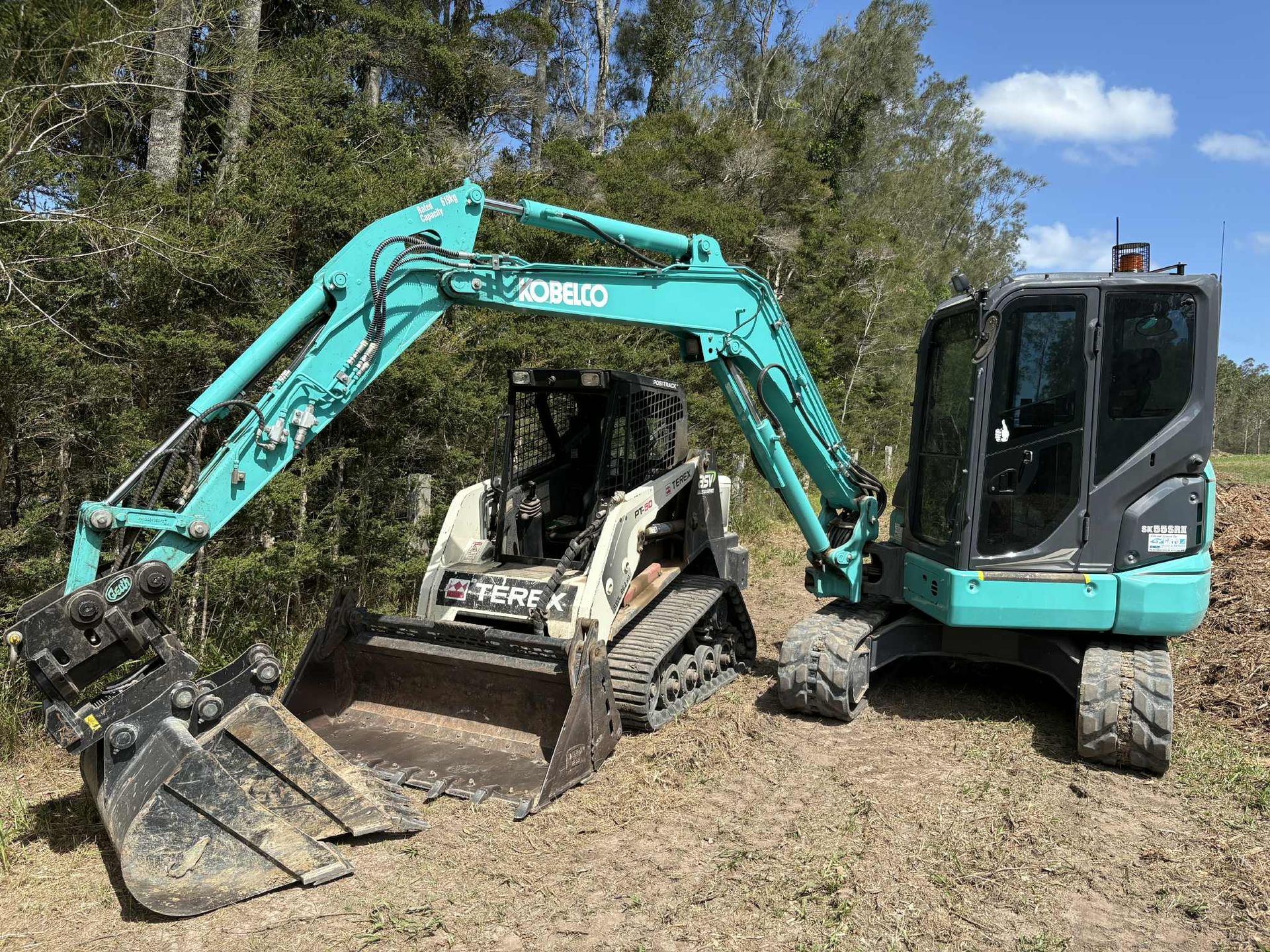 A teal excavator and a white skid steer loader parked side-by-side in a wooded area.