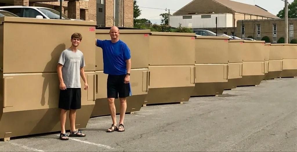 Two men are standing in front of a row of large cardboard boxes.