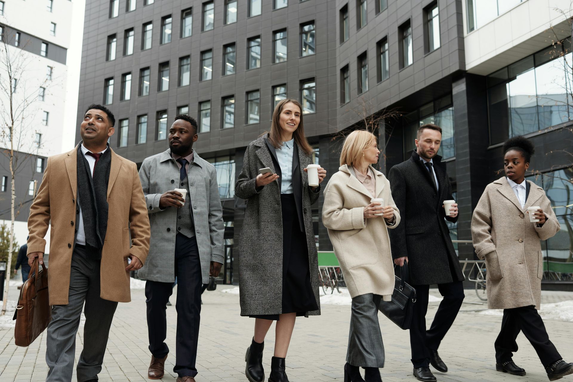 Six people walking on a city sidewalk beside a modern office building, dressed in winter coats.