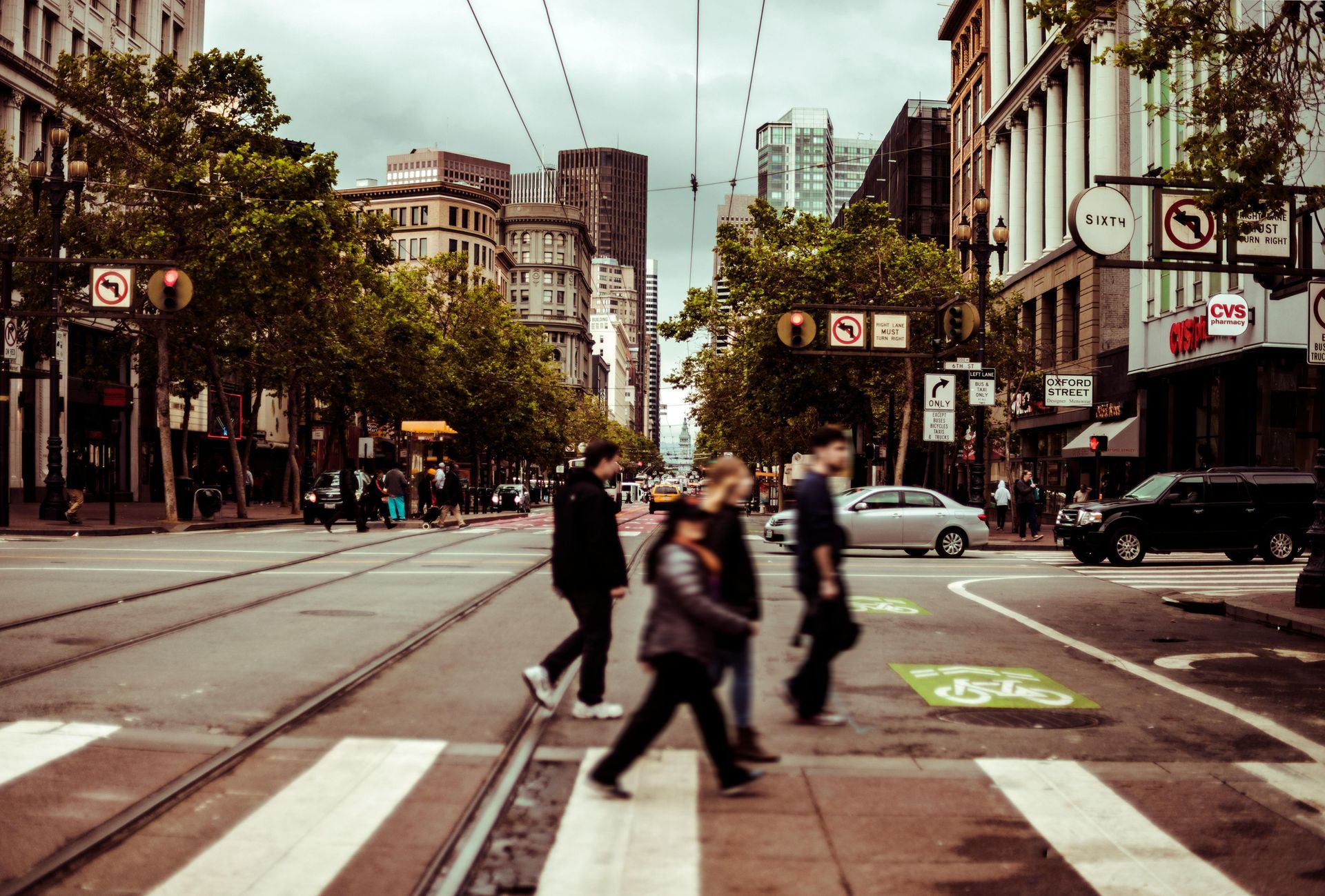 Pedestrians crossing a city street at a crosswalk, with buildings, trees, and cable car tracks in the background