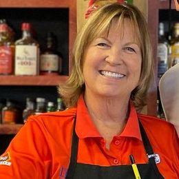 a woman in a red shirt and apron is smiling in a liquor store.