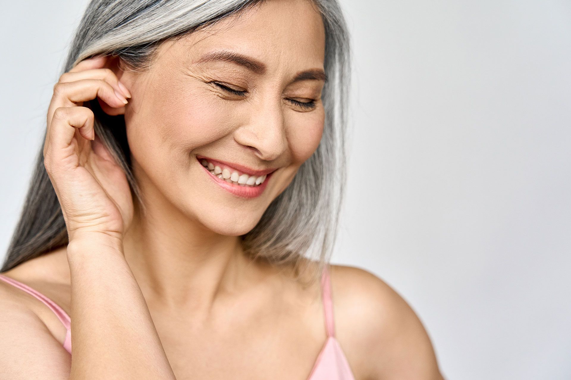 Woman with gray hair and closed eyes smiles, hand touching ear, wearing pink tank top.