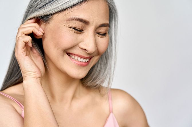 Woman with gray hair and closed eyes smiles, hand touching ear, wearing pink tank top.