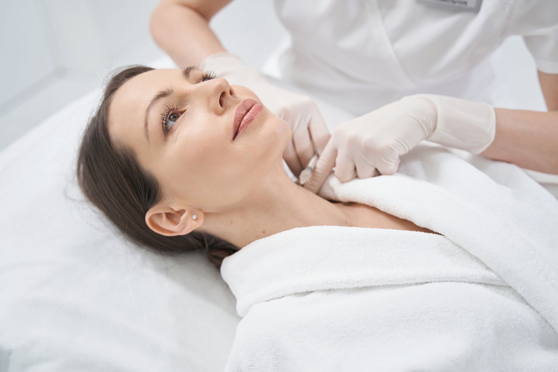 Woman receiving cosmetic treatment on her neck. She lies on a bed in a white robe.