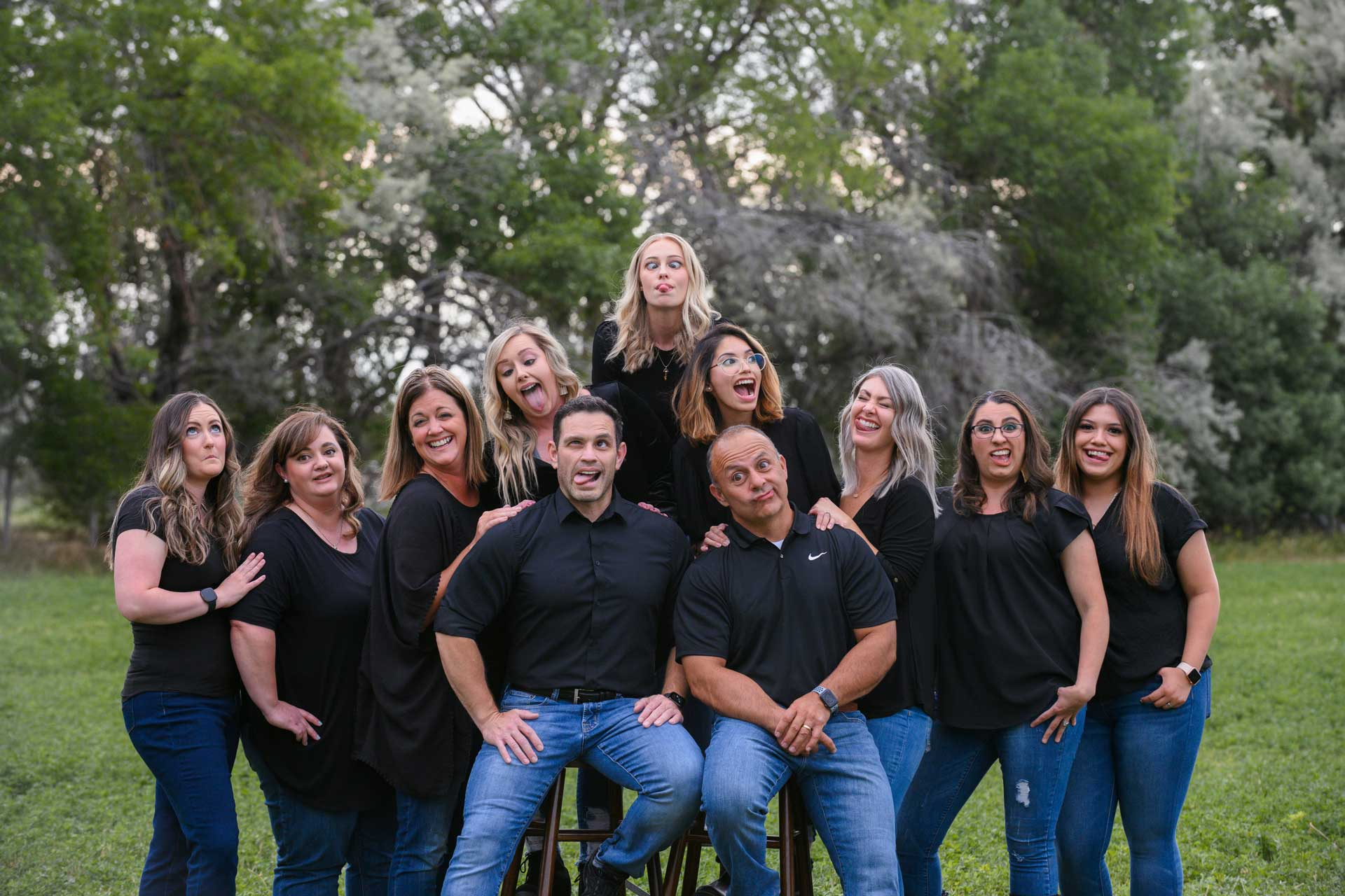 Group of people, mostly women, making silly faces; posing outside near trees, wearing black tops and jeans.