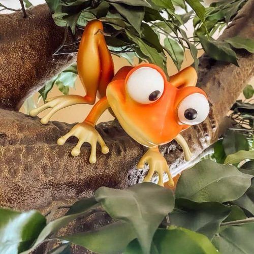 Orange cartoon frog with oversized eyes perched on a brown branch with green leaves.