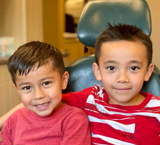 Two young boys smiling, one in red shirt, other in striped shirt, seated in a dental chair.