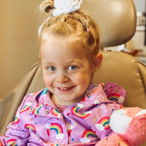 Girl with blonde hair in a pink rainbow jacket smiles in a dental chair, holding a stuffed animal.