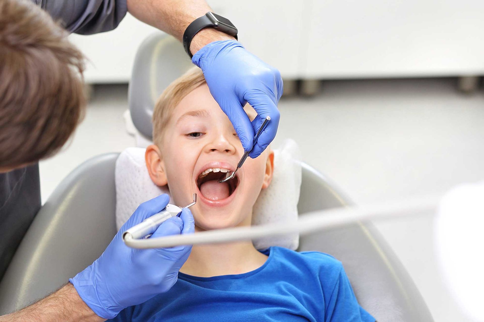 Child in dental chair, mouth open, dentist examining teeth with tools. Blue gloves, bright setting.
