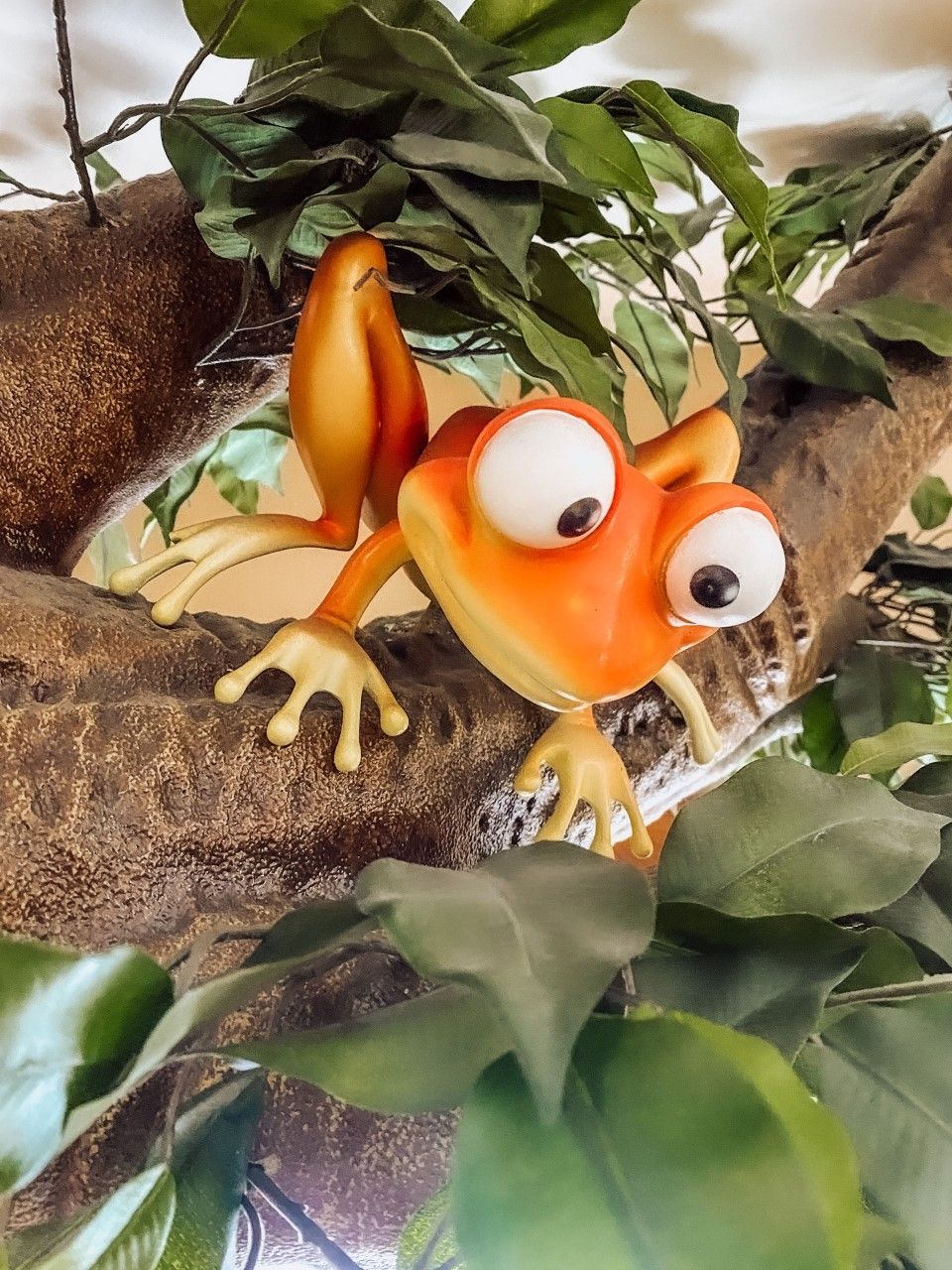 Orange cartoon frog with large eyes on a tree branch, surrounded by green leaves.