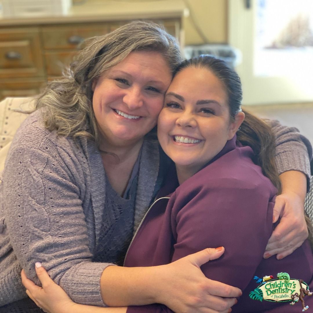 Two smiling women hugging closely indoors. One in purple scrubs, the other in a gray sweater.