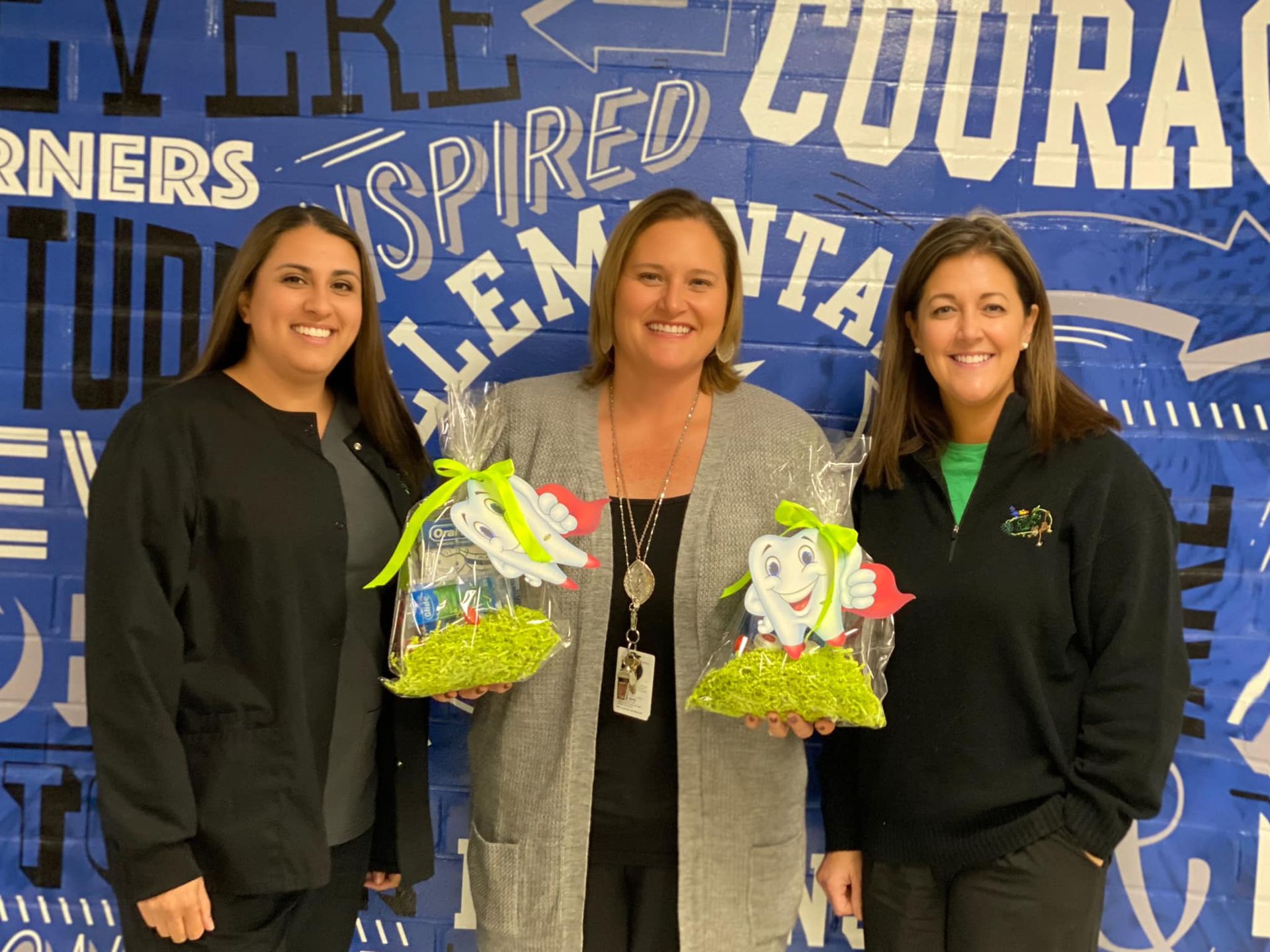 Three women hold gift baskets with tooth decorations in front of a blue school backdrop.