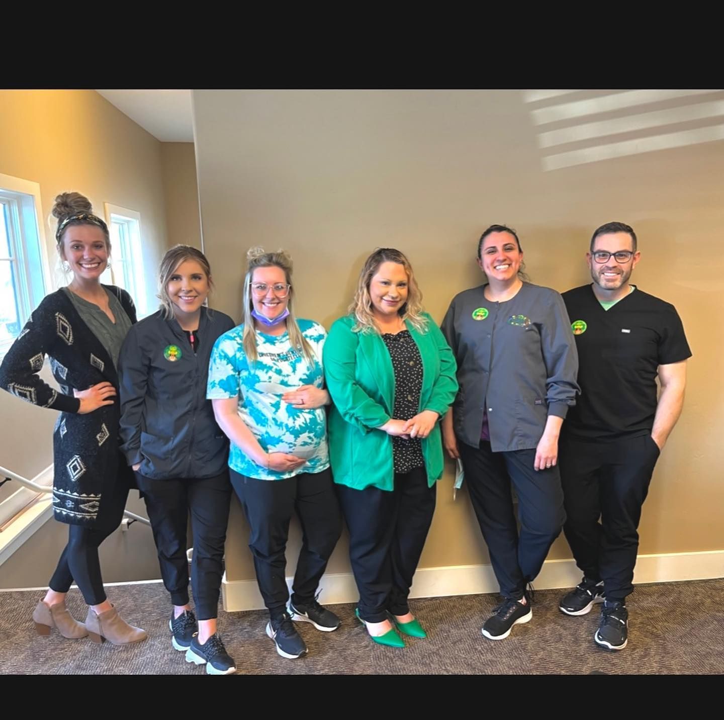Six people in a group posing. They are standing near a beige wall, a window, and stairs. Some wear scrubs.