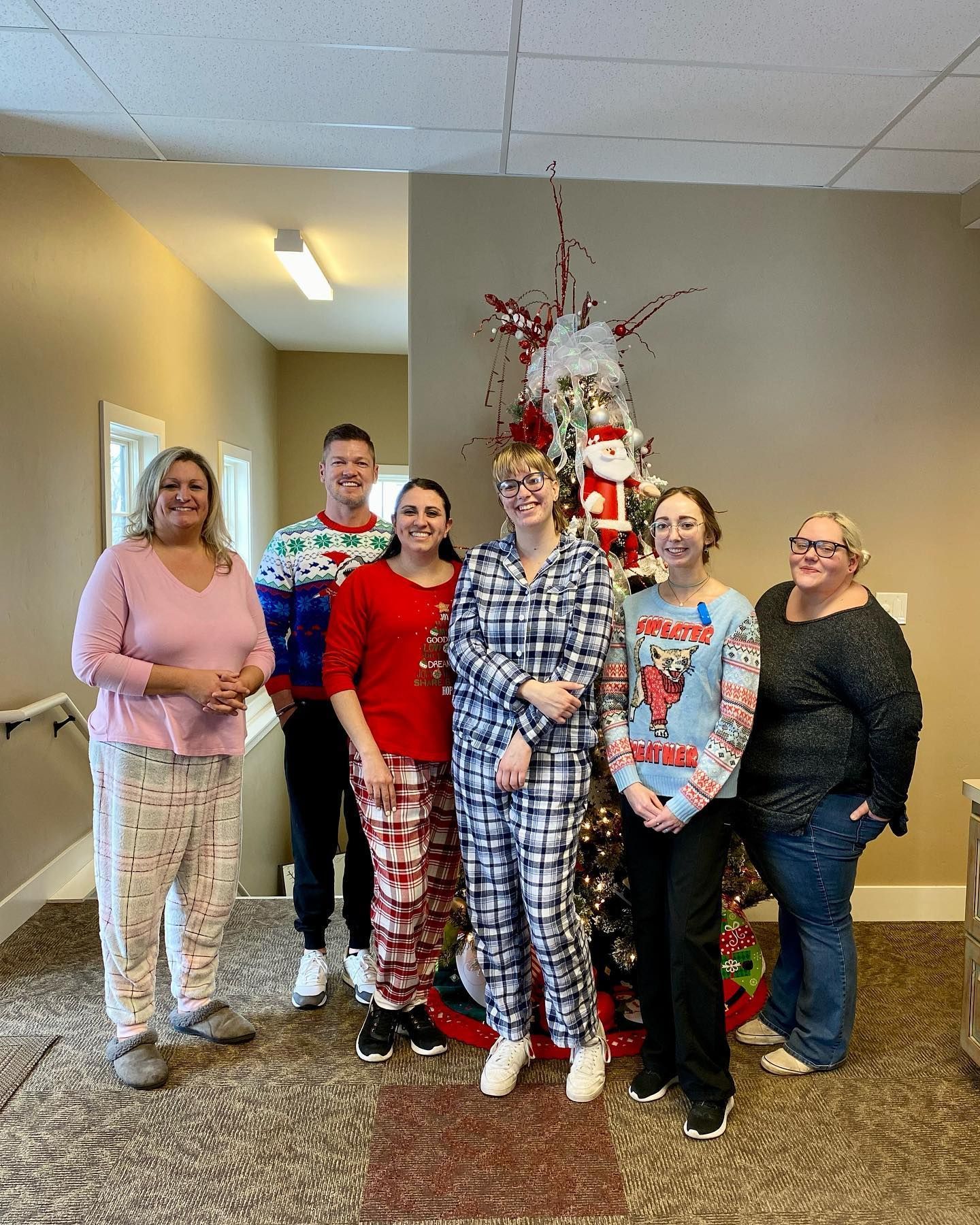 People in pajamas and festive sweaters pose in front of a decorated Christmas tree indoors.