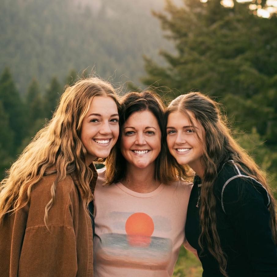 Woman and two young women smiling, posed in front of trees.