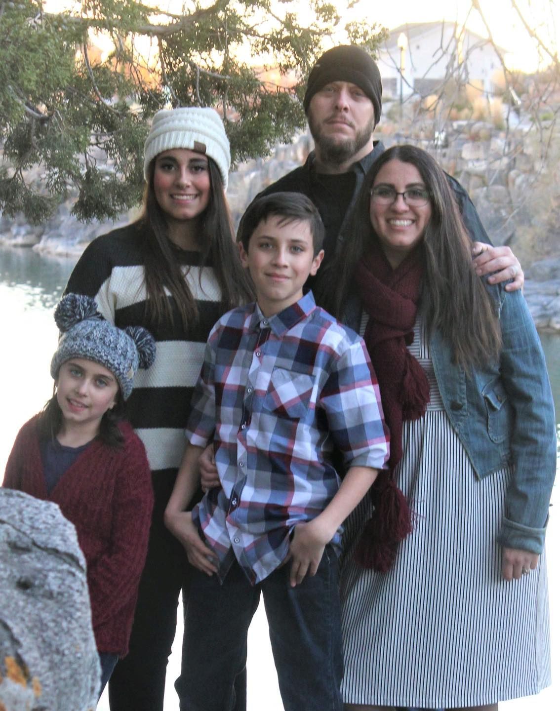 Family of five poses outdoors near water. Adults wear beanies; the boy in a plaid shirt.