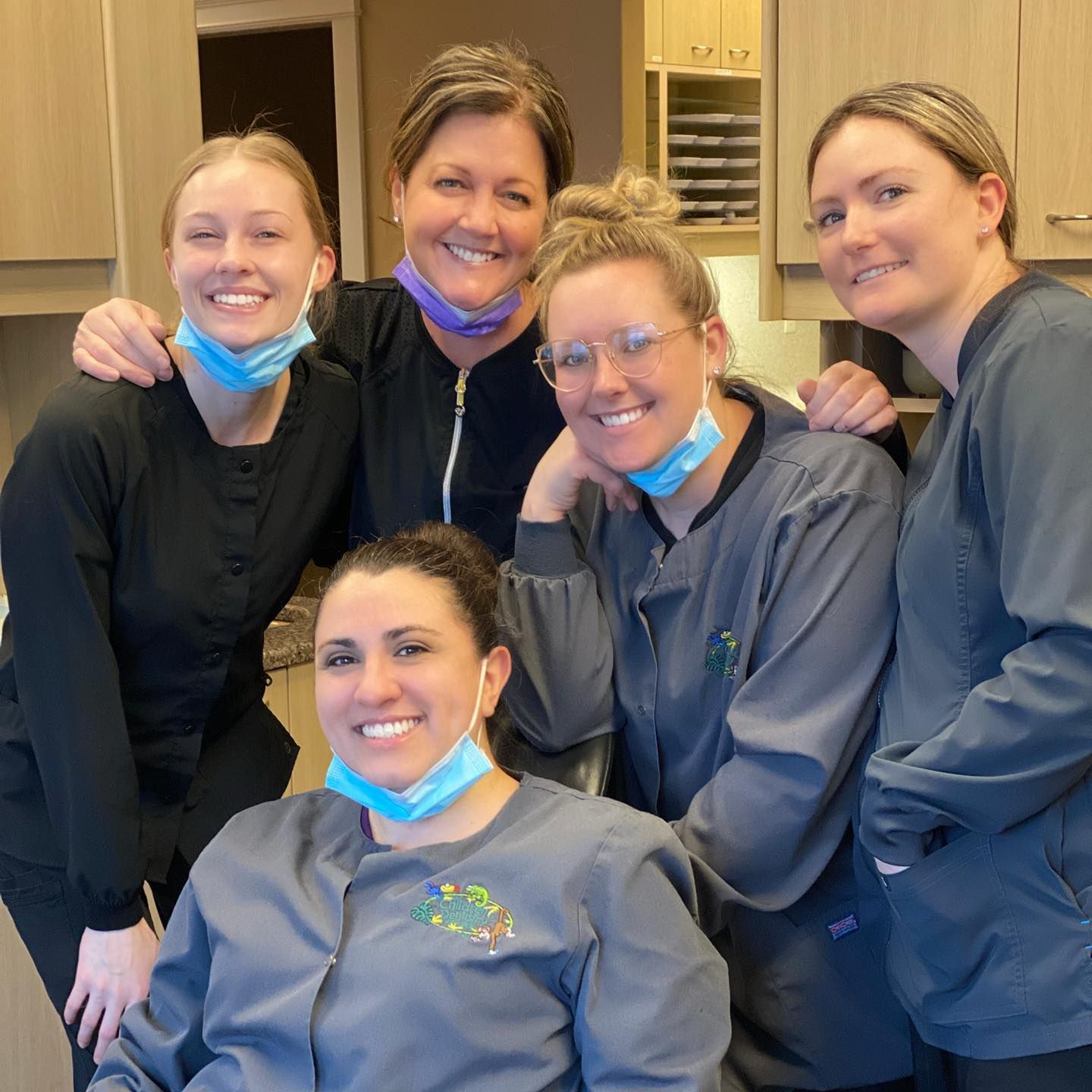 Five dental professionals smiling, some wearing masks and scrubs, in an office setting.