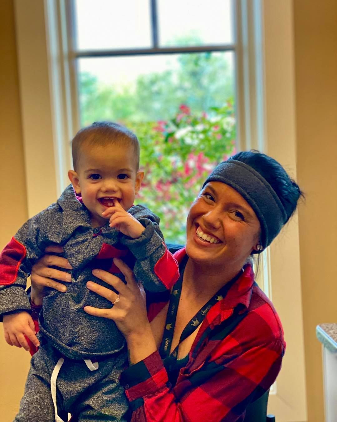 Woman holding a smiling child; both are near a window. Woman wears a red plaid shirt, child in gray sweatsuit.