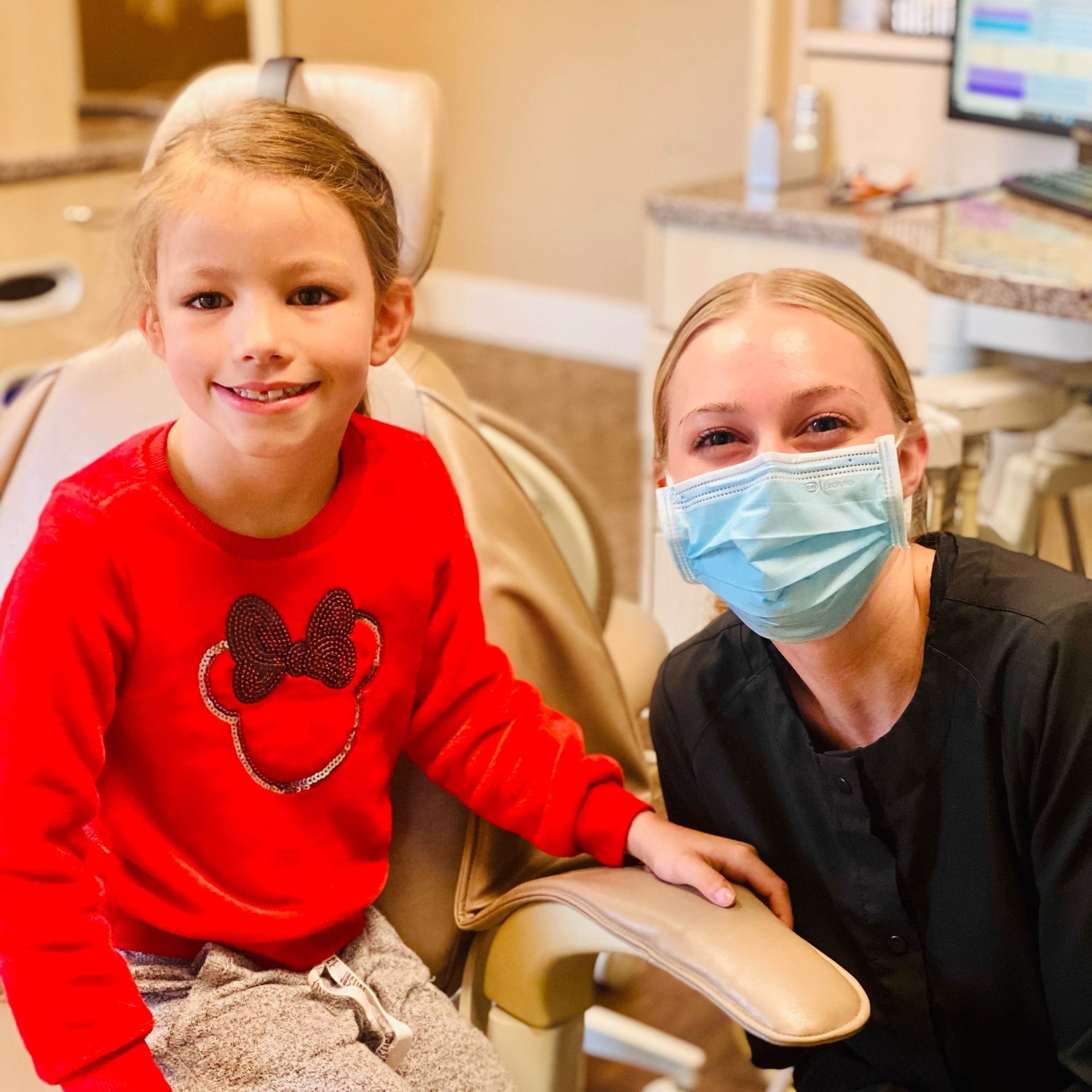 Girl smiles in dentist chair next to a masked dental assistant. Both are in an office setting.