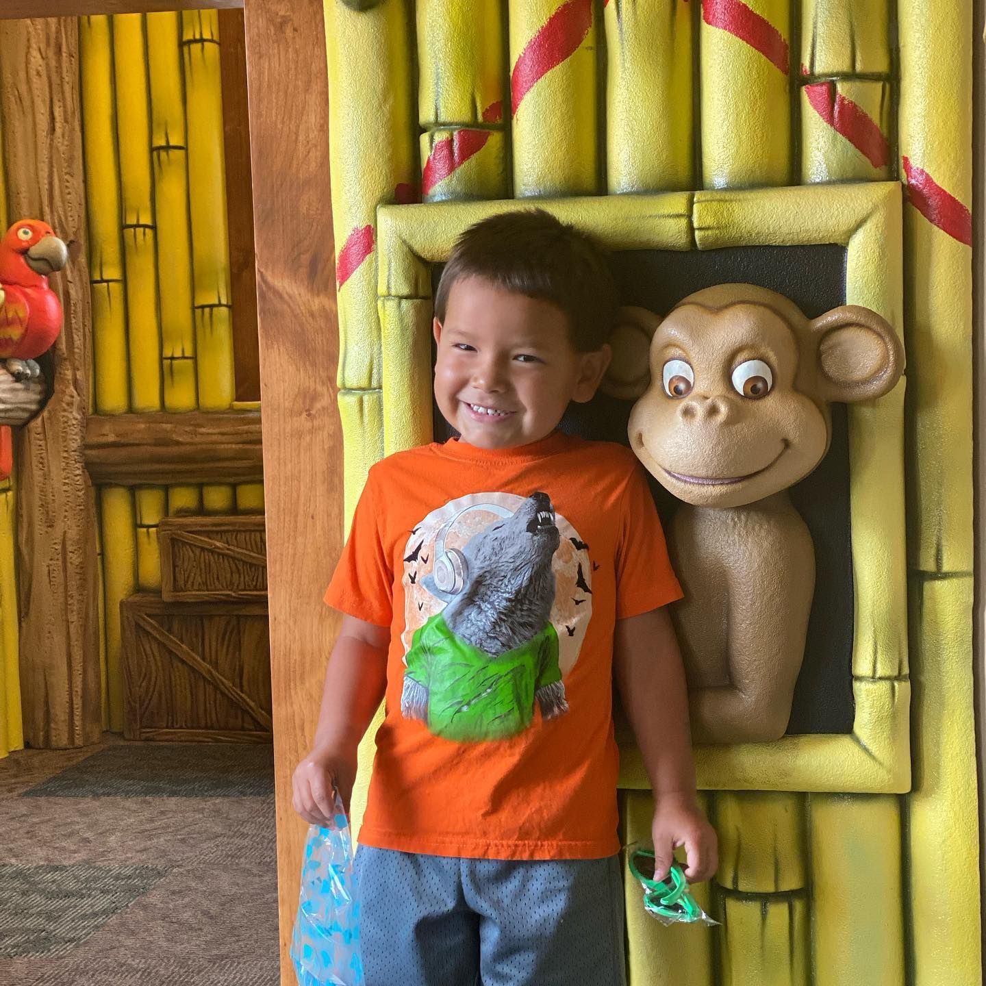 Smiling child in orange shirt stands by a monkey statue in a jungle-themed setting.