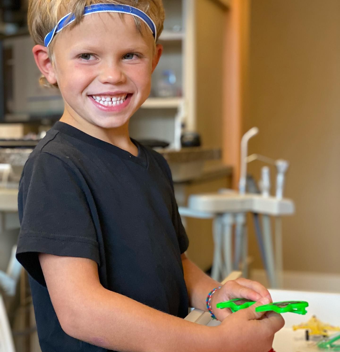 Boy wearing headband smiling, holding green toy airplane. Kitchen background.