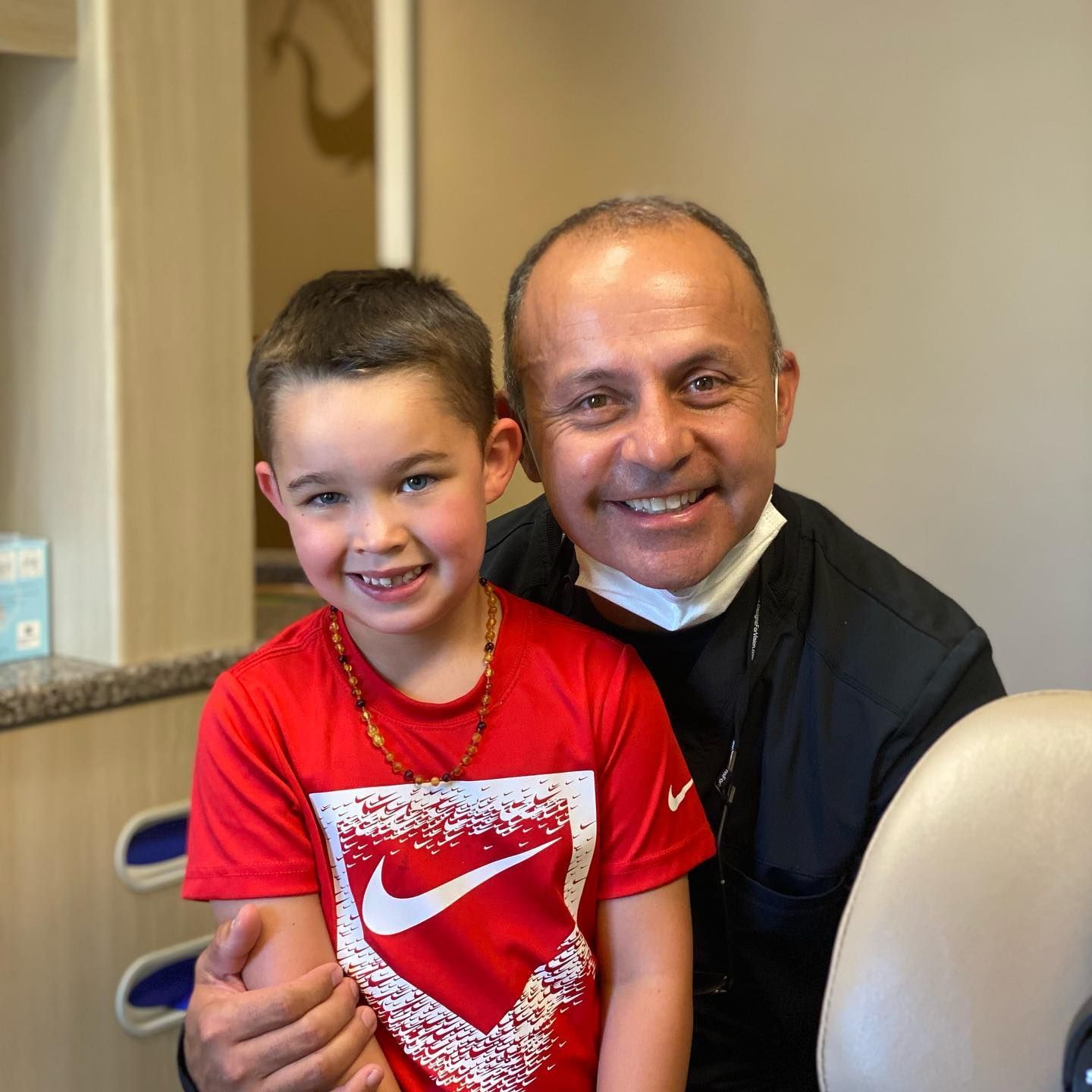 A smiling child in a red shirt with a Nike logo and a dentist, both smiling in an office setting.