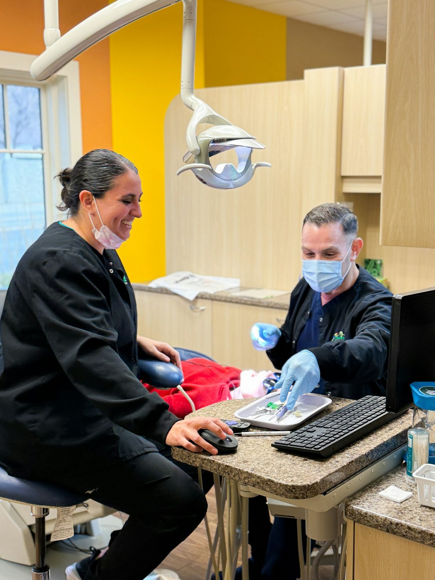 Dental office: Two dental professionals; one with mask selects tools, the other operates computer, smiling.