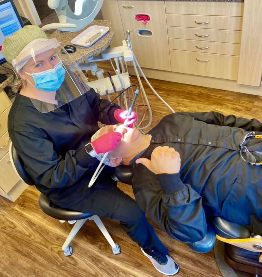 Dentist in scrubs and face shield examining a patient's teeth in a dental office.