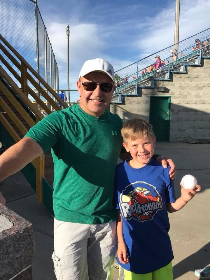 Man and boy at a baseball field; boy holds a ball. Bright colors, sunny sky.