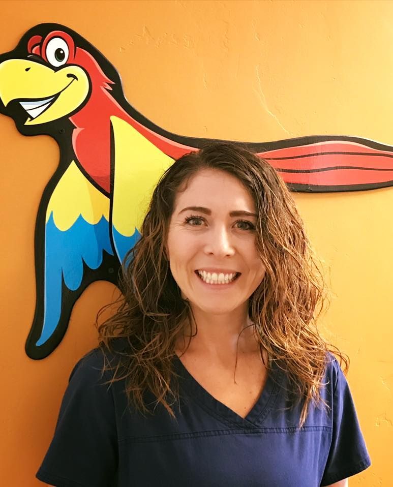 Woman in blue scrubs smiles in front of a colorful parrot logo on orange wall.