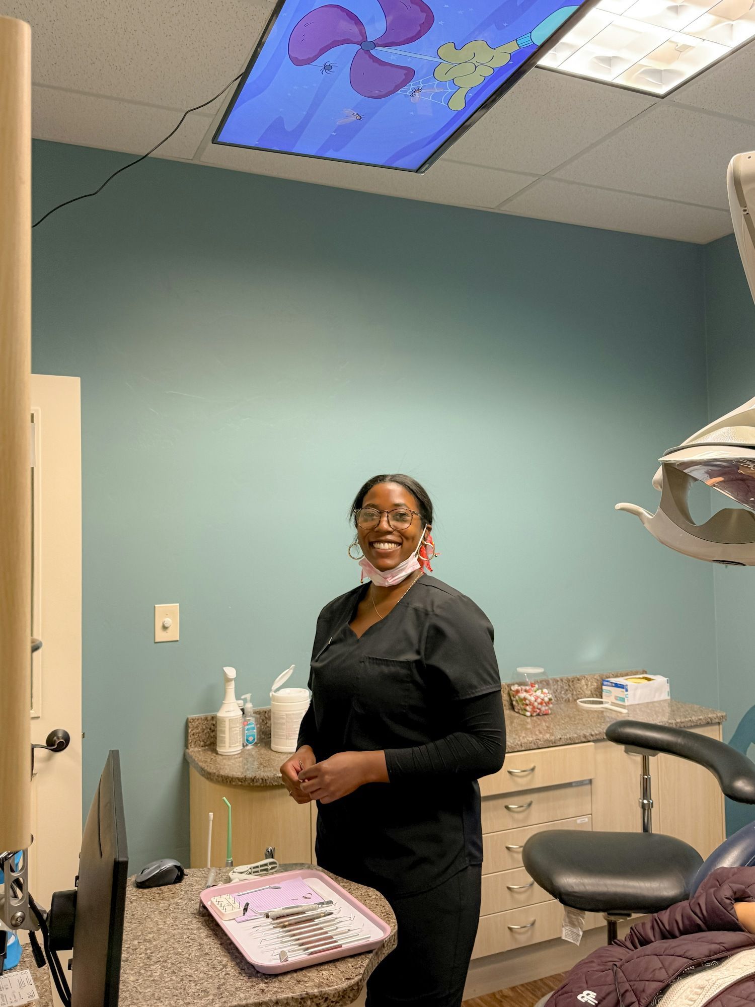 Dental hygienist in scrubs smiles in a dentist's office with cartoon ceiling art.