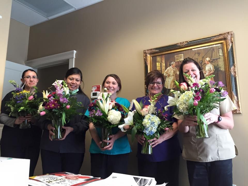 Five people holding bouquets of flowers, smiling in an office setting with a painting on the wall.